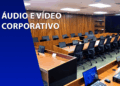 Sala de conferências moderna com bancadas de madeira, cadeiras pretas e sistemas multimídia integrados. À esquerda, um bloco azul com o texto "ÁUDIO E VÍDEO CORPORATIVO".