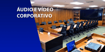 Sala de conferências moderna com bancadas de madeira, cadeiras pretas e sistemas multimídia integrados. À esquerda, um bloco azul com o texto "ÁUDIO E VÍDEO CORPORATIVO".