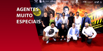 Elenco e equipe do filme "Agentes Muito Especiais" reunidos em um tapete vermelho à frente de um painel promocional com explosões e o rosto de Paulo Gustavo ao fundo. À esquerda, uma faixa vermelha com o título da comédia policial nacional.