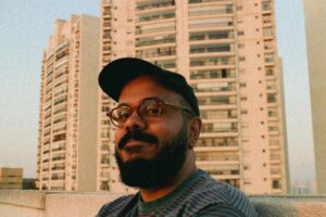 etrato do autor Caio Martim em primeiro plano. Ele usa óculos de grau redondos e boné preto, posicionado à frente de prédios residenciais sob a luz do pôr do sol.
