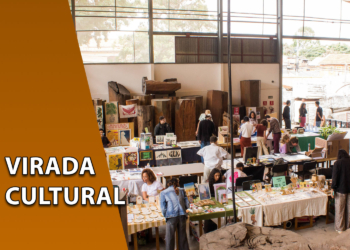 Visão ampla de um galpão industrial iluminado com diversas mesas de exposição de artesanato e público circulando. À esquerda, uma faixa marrom inclinada com o texto "VIRADA CULTURAL".