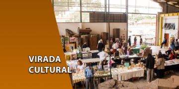 Visão ampla de um galpão industrial iluminado com diversas mesas de exposição de artesanato e público circulando. À esquerda, uma faixa marrom inclinada com o texto "VIRADA CULTURAL".