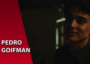 Retrato em baixa luminosidade do ator Pedro Goifman sorrindo levemente. À esquerda, uma faixa vermelha inclinada com o texto "PEDRO GOIFMAN".