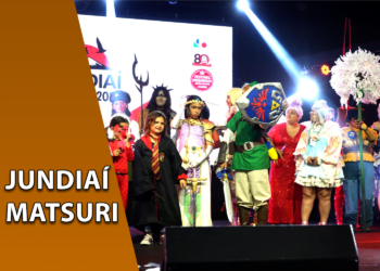 Diversos cosplayers reunidos no palco de um evento de cultura japonesa sob luzes de refletores. À esquerda, uma faixa marrom inclinada com o texto "JUNDIAÍ MATSURI".
