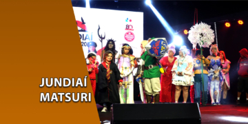Diversos cosplayers reunidos no palco de um evento de cultura japonesa sob luzes de refletores. À esquerda, uma faixa marrom inclinada com o texto "JUNDIAÍ MATSURI".