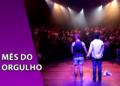 Dois atores de costas e de mãos dadas no palco de um teatro iluminado por holofotes, diante de uma plateia aplaudindo. À esquerda, uma faixa roxa inclinada com o texto "MÊS DO ORGULHO".