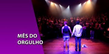 Dois atores de costas e de mãos dadas no palco de um teatro iluminado por holofotes, diante de uma plateia aplaudindo. À esquerda, uma faixa roxa inclinada com o texto "MÊS DO ORGULHO".