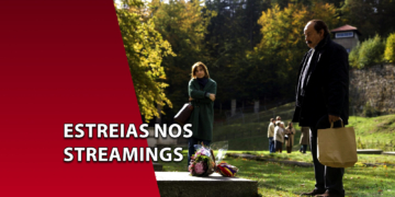 Homem de casaco escuro segurando uma sacola de papel em um gramado ao ar livre, olhando para um túmulo com flores, enquanto uma mulher o observa ao fundo. À esquerda, uma faixa vermelha inclinada com o texto "ESTREIAS NOS STREAMINGS".