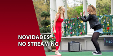 Duas mulheres em cima de um palco ao ar livre gesticulando de forma expressiva e cômica diante de um amplificador. À esquerda, uma faixa vermelha inclinada com o texto "NOVIDADES NO STREAMING".