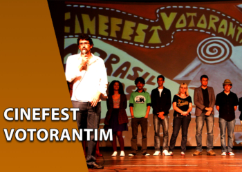 Um homem de camisa branca falando ao microfone no centro de um palco, ao lado de um grupo de pessoas alinhadas em frente a um grande painel pintado com o nome "CINEFEST VOTORANTIM". À esquerda, uma faixa marrom inclinada com o texto "CINEFEST VOTORANTIM".