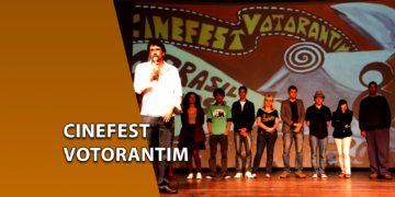 Um homem de camisa branca falando ao microfone no centro de um palco, ao lado de um grupo de pessoas alinhadas em frente a um grande painel pintado com o nome "CINEFEST VOTORANTIM". À esquerda, uma faixa marrom inclinada com o texto "CINEFEST VOTORANTIM".