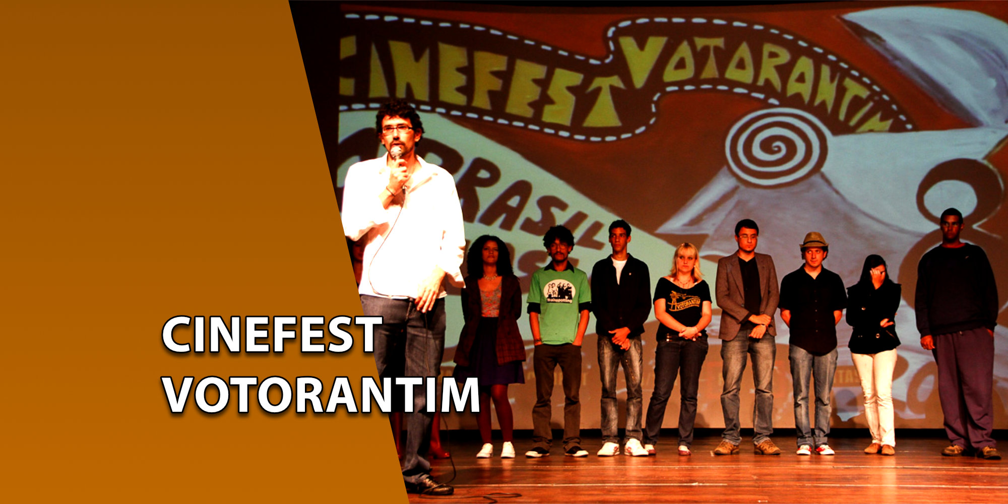 Um homem de camisa branca falando ao microfone no centro de um palco, ao lado de um grupo de pessoas alinhadas em frente a um grande painel pintado com o nome "CINEFEST VOTORANTIM". À esquerda, uma faixa marrom inclinada com o texto "CINEFEST VOTORANTIM".