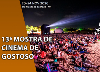 Plateia sentada em espreguiçadeiras na areia da praia assistindo a uma projeção noturna ao ar livre durante a Mostra de Cinema de Gostoso. À esquerda, uma faixa marrom inclinada com o texto "13ª MOSTRA DE CINEMA DE GOSTOSO".